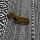 Fallenbrokenstreet brass bottle opener photographed on textured fabric. Compact 5cm x 2cm design with embossed Fallenbrokenstreet logo, crafted from solid brass. A timeless accessory for festivals, travel, and home.