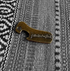 Fallenbrokenstreet brass bottle opener photographed on textured fabric. Compact 5cm x 2cm design with embossed Fallenbrokenstreet logo, crafted from solid brass. A timeless accessory for festivals, travel, and home.