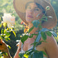 Woman wears the Fallenbrokenstreet straw hat in a lush garden, surrounded by blooming leaves and dappled light. The hat’s feather and ribbon details add vintage charm.

