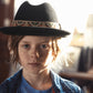 Child wearing a black fedora with a patterned band, standing in a softly lit room.