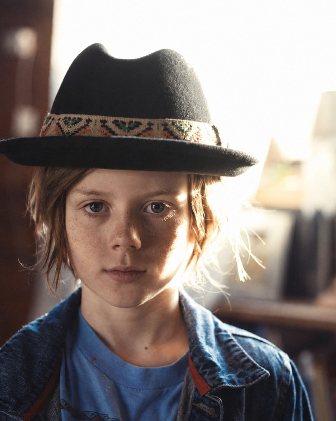 Child wearing a black fedora with a patterned band, standing in a softly lit room.