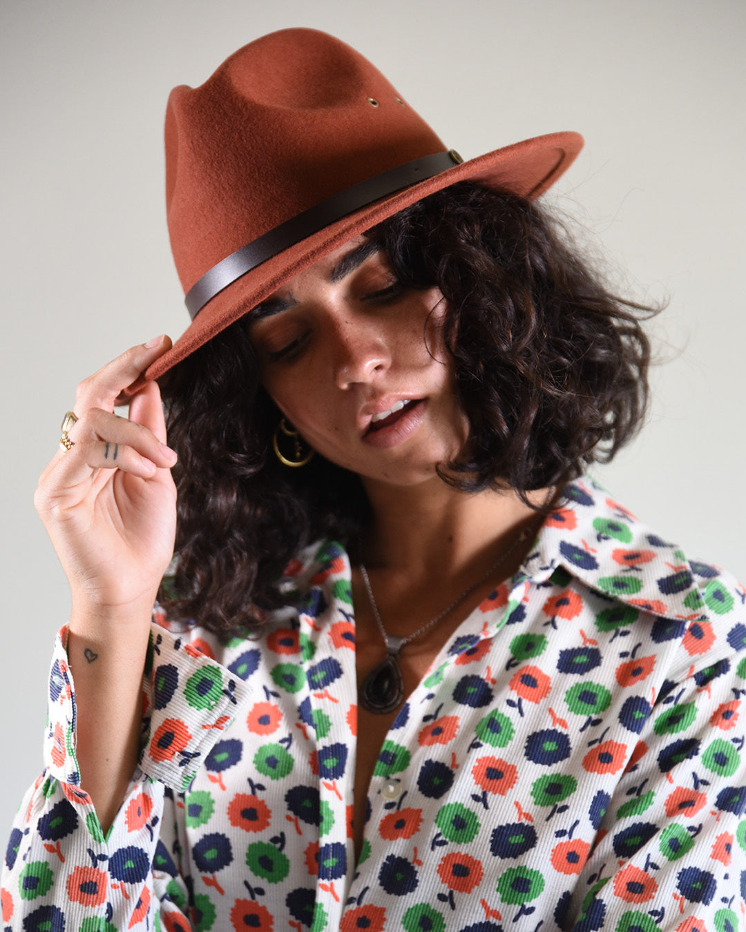 Woman wearing a brown fedora hat and a colorful floral shirt against a plain background