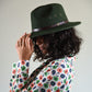 Person wearing a green fedora hat with a brown band against a plain background