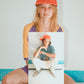 Woman in orange and purple tee sitting cross-legged, holding framed photo of the cap.

