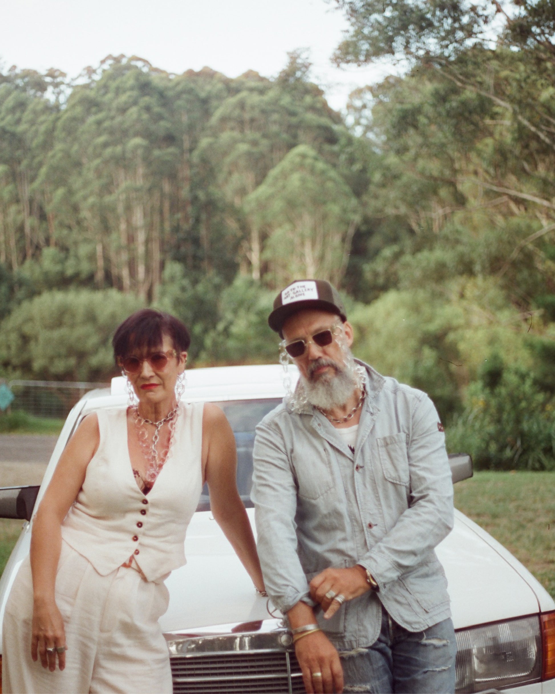 Couple seated outdoors wearing matching sunglasses and neutral-toned outfits. Captures a relaxed, stylish moment in natural light.

