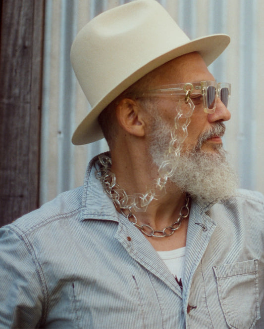 Older male model wears The Mischief – Skywalker/Amber sunglasses with white hat and button-up shirt. Styled in natural light with a relaxed, vintage feel.

