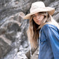 Model perched on rocky terrain wearing The Lover Straw Hat – Natural. This shot highlights the hat’s flexibility and comfort in real-world adventures, from hikes to beachside wandering. The natural tones blend beautifully with the rugged backdrop.

