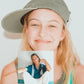 Close-up of smiling model wearing dad cap and holding instant photo of herself and friend.

